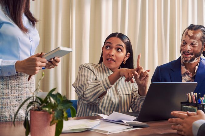 A Deaf Latina Puerto Rican woman communicates in American Sign Language to a caucasian woman with long straight brown hair wearing business attire standing next to her while seated at a conference room table with two colleagues. The Deaf woman has long deep brown hair and is wearing a striped business shirt and hoop earrings. An African American man with olive and porcelain skin, Non-Segmental Vitiligo, and brown barrel-twist dreadlocks looks at the Deaf woman with focus. A laptop, business planning documents, a cup of pens, and a potted plant are on the conference room table.