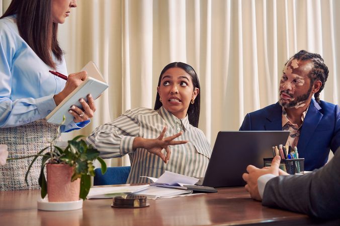 A Deaf Latina Puerto Rican woman communicates in American Sign Language to a caucasian woman with long straight brown hair wearing business attire standing next to her while seated at a conference room table with two colleagues. The Deaf woman has long deep brown hair and wears a striped business shirt and hoop earrings. An African American man with olive and porcelain skin, Non-Segmental Vitiligo, and brown barrel-twist dreadlocks wearing a business suit looks at the Deaf woman with focus. A laptop, business planning documents, a cup of pens, and a potted plant are on the conference room table.