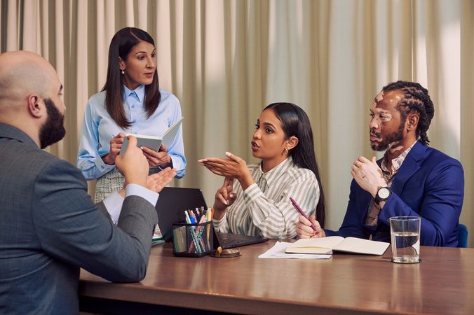 A bald white hard of hearing sign language interpreter and a Deaf Latina Puerto Rican woman communicate in American Sign Language at a conference room table. The interpreter is wearing a gray business suit. The Deaf woman has long deep brown hair and is wearing a striped business shirt and hoop earrings. An African American man with olive and porcelain skin, Non-Segmental Vitiligo, and brown barrel-twist dreadlocks wearing a blue business suit looks across the table. A caucasian woman with long straight brown hair wearing business attire stands next to the Deaf woman, holding a notebook and pen. A laptop, business planning documents, a cup of pens, and a glass of water are on the conference room table.