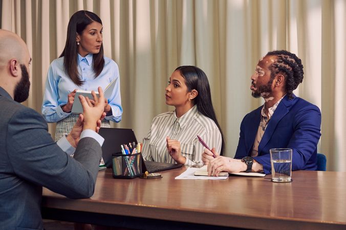 A caucasian woman with long straight brown hair wearing business attire speaks to a Deaf Latina Puerto Rican woman sitting next to her at a conference room table. The Deaf woman watches a bald white hard of hearing sign language interpreter translate the CaucAsian woman's speech into American Sign Language. The Deaf woman has long deep brown hair and is wearing a striped business shirt and hoop earrings. The interpreter is wearing a gray business suit. An African American man with olive and porcelain skin and Non-Segmental Vitiligo looks at the CaucAsian woman. The man has brown barrel-twist dreadlocks and is wearing a blue business suit. A laptop, business planning documents, a cup of pens, and a potted plant are on the conference room table.