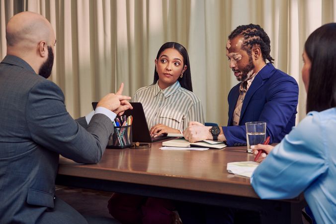 A bald white hard of hearing sign language interpreter translates speech into American Sign Language for a Deaf Latina Puerto Rican woman sitting across from him at a conference room table. An African American man with olive and porcelain skin and Non-Segmental Vitiligo is seated next to her at the table, writing in a notebook. A caucasian woman with long brown hair writes in a notebook across the table. All four colleagues are wearing business attire.