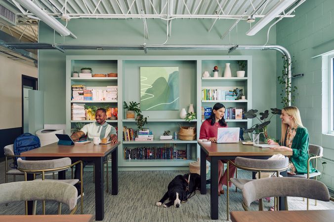 Colleagues chat and work in a modern coworking office space. At one table, a young white woman with partially paralyzed arms and hands chats with a young pale white blind woman. A black and white guide dog with a guide harness lies on the floor next to the blind woman, who is typing on a laptop computer with headphones nearby. She has shiny black hair styled in waves and is wearing a chunky knitted pink sweater and dusky pink flared pants. The white woman with partially paralyzed arms and hands is smiling while writing in a notebook, with a glass of water with a straw in it nearby. She has long, straight blonde hair and is wearing a floral dress and a green blazer. At another table, a Black man smiles while looking at his tablet computer. He is wearing a white and green polo shirt and a pastry and mug are nearby. The coworking office space is painted a warm mint green and has a large bookcase displaying several books, plants, art pieces, and clay vases.