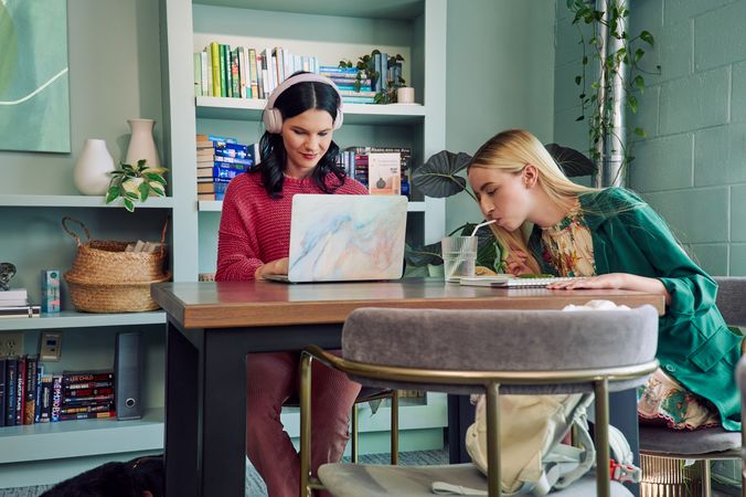 A young white woman with long, straight blonde hair and partially paralyzed arms and hands bends forward to drink water from a cup through a straw. She is sitting at a table in a modern coworking office space with a young pale white blind woman, who is typing on a laptop computer. The blind woman has shiny black hair styled in waves and is wearing over the ear headphones, a chunky knitted pink sweater, and dusky pink flared pants. The woman with partially paralyzed arms and hands is wearing a floral dress with pink trim and a green blazer. The coworking office space is painted a warm mint green and has a large bookcase and features several books, plants, a rattan basket, and clay vases.