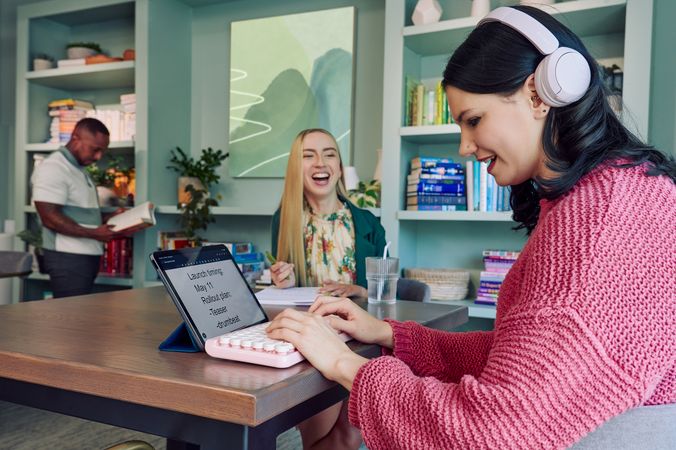 A young pale white blind woman is sitting at a table, typing on a keyboard connected to a tablet computer in a modern coworking office space. Visible on her tablet is a document in large print that reads "Launch timing: May 11. Rollout plan: Teaser. Drumbeat.\" She is smiling as she types. Across from her at the table, a young white woman with partially paralyzed arms and hands laughs with the blind woman as she holds a pen to a notebook in front of her. A glass of water with a straw in it is nearby. The woman with partially paralyzed arms and hands has long straight blonde hair and is wearing a floral dress and green blazer. The blind woman has shiny black hair styled in waves and is wearing over the ear headphones that partially cover one ear and a chunky knitted pink sweater. The coworking office space is painted a warm mint green and features a large bookcase with several books, plants, and a large abstract art piece.