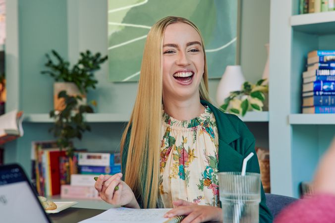A young white woman with long, straight blonde hair and partially paralyzed arms and hands laughs with a wide smile. She is sitting at a table in a modern coworking office space holding a pen with an open notebook in front of her. A glass of water with a straw in it is nearby. She is wearing a floral dress and a green blazer. The coworking office space is painted a warm mint green and features a large bookcase with several books, plants, and a large abstract art piece.