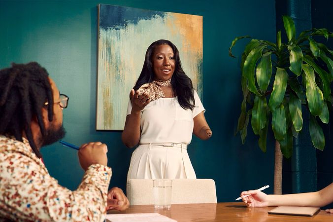 An executive leads a team meeting standing in a dark conference room, using her arms as she speaks. The executive is a Black American woman and below-elbow congenital amputee wearing a cream blouse and pants, with a patterned scarf around her neck. Her hair is side parted and coiffed. A neurodivergent Black male sitting at the conference table looks at the executive while holding a pen. He has a notebook in front of him. He has locs and is wearing glasses, a floral print button down shirt, and a red tie. The conference room is painted dark teal and features an abstract art piece and a large potted plant.