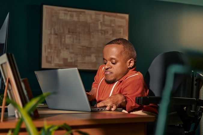 A black man who is a little person is seated in his power wheelchair in a dark teal home office. He types on a laptop on his desk, with a monitor and framed photos nearby.  An abstract art piece is on the wall behind him. He has closely cropped black hair, brown eyes, and is wearing a rust-orange knit quarter-zip sweater with white vertical stripes, light khaki trousers, and a small stud earring.