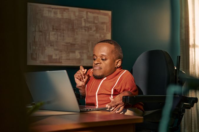 A black man who is a little person is seated in his power wheelchair in a dark teal home office. He types on a laptop on his desk, his hand held up in concentration.  An abstract art piece is on the wall behind him. He has closely cropped black hair, brown eyes, and is wearing a rust-orange knit quarter-zip sweater with white vertical stripes, light khaki trousers, and a small stud earring.