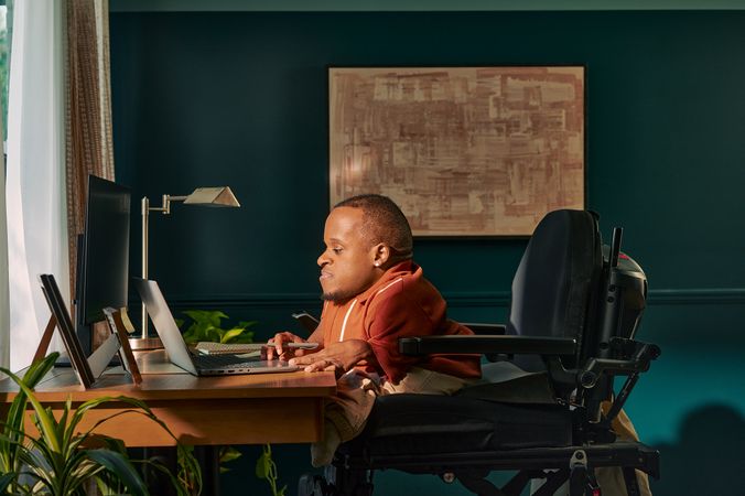 A black man who is a little person is seated in his power wheelchair in a dark teal home office. He holds a pen and types on his laptop. A monitor, notebook, pen, lamp, and framed photos are on the desk in front of him. His wheelchair is in an elevated position, allowing him to be level with his desk. He is facing a window, letting in light through white and patterned curtains. His desk is surrounded by several house plants and an abstract art piece. He has closely cropped black hair, brown eyes, and is wearing a rust-orange knit quarter-zip sweater with white vertical stripes, light khaki trousers, and a small stud earring. A tote bag hangs on the back of his wheelchair.