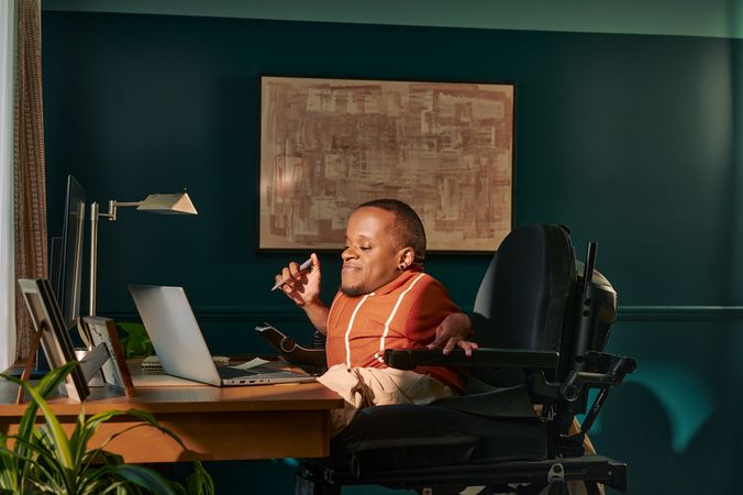 A black man who is a little person is seated in his power wheelchair in a dark teal home office. He holds a pen and looks down at his laptop. A monitor, notebook, pen, lamp, and framed photos are on the desk in front of him. His desk is surrounded by house plants and an abstract art piece. He has closely cropped black hair, brown eyes, and is wearing a rust-orange knit quarter-zip sweater with white vertical stripes, light khaki trousers, and a small stud earring. A tote bag hangs on the back of his wheelchair.
