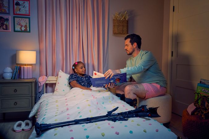A white man with Cerebral Palsy who wears leg braces shows a children's book titled "Extraordinary Jordyn and Her Bionic Ears" to his daughter in her bedroom at night. His daughter is a Black multiracial American girl with thick braided red hair adorned with two flower barrettes. She is tucked into bed and is wearing dark blue patterned pajamas and looks at the open book. The father's black forearm crutches lie on the foot of the bed. He has dark brown curly hair, short brown facial hair, and is wearing a light green sweater with pink shorts. He has a tattoo of the Roman numeral two followed by a slash and the number three. The child's room is painted light pink and includes a nightstand, lamp, a pair of slippers, curtains, and children's books. Her bedspread is white with blue, pink, and yellow polka dots. Framed illustrations of Black women hang on the wall.