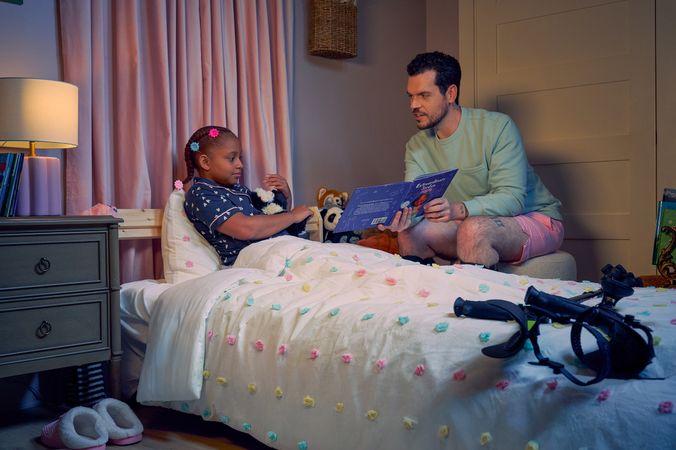 A white man with Cerebral Palsy who wears leg braces holds a children's book titled "Extraordinary Jordyn and Her Bionic Ears" to his daughter in her bedroom at night. His daughter is a Black multiracial American girl with thick braided red hair adorned with two flower barrettes and is wearing blue patterned pajamas. She is tucked into bed, holding a plushie animal and looks at the open book. The father's black forearm crutches lie on the foot of the bed. He has dark brown curly hair, short brown facial hair, and is wearing a light green sweater with pink shorts. He has a tattoo of the Roman numeral two followed by a slash and the number three. The child's room is painted light pink and includes a nightstand, lamp, a pair of slippers, curtains, and children's books. Her bedspread is white with blue, pink, and yellow polka dots. Framed illustrations of Black women hang on the wall.