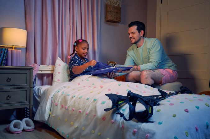 A white man with Cerebral Palsy who wears leg braces holds a children's book titled "Extraordinary Jordyn and Her Bionic Ears" to his daughter in her bedroom at night. His daughter is a Black multiracial American girl with thick braided red hair adorned with two flower barrettes and is wearing blue patterned pajamas. She is tucked into bed, holding a plushie animal and turns a page in the book. The father's black forearm crutches lie on the foot of the bed. He has dark brown curly hair, short brown facial hair, and is wearing a light green sweater with pink shorts. He has a tattoo of the Roman numeral two followed by a slash and the number three. The child's room is painted light pink and includes a nightstand, lamp, a pair of slippers, curtains, and children's books. Her bedspread is white with blue, pink, and yellow polka dots. Framed illustrations of Black women hang on the wall.
