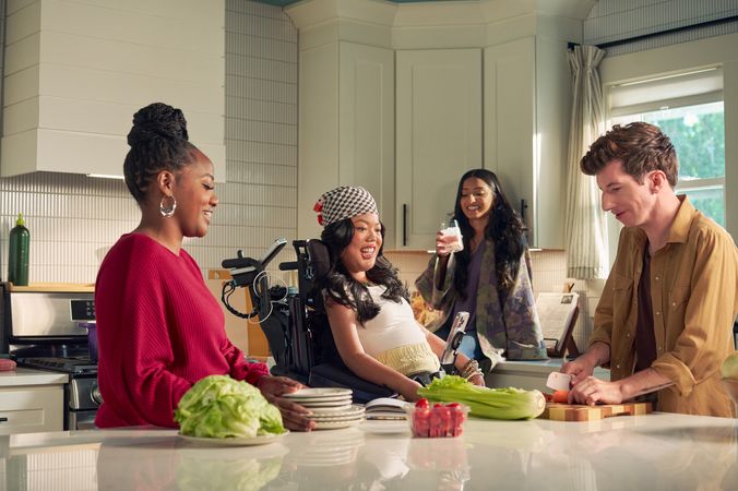 A SouthEast Asian woman with curled black hair seated in her power wheelchair smiles and watches her roommate chop vegetables in their bright kitchen. Her smart phone is mounted to her wheelchair. Her roommate is a white male with Moebius Syndrome and a face that is paralyzed. The SouthEast Asian woman is wearing a white top with striped, yellow linen pants and a checkered headscarf. He has short, wavy, reddish brown hair and is wearing a tan button down shirt with the sleeves rolled up, showing arm tattoos. Two other roommates laugh and hang out nearby. One roommate has long dark hair and is wearing a purple floral dress, the other is a black female with a braided bun and maroon top.