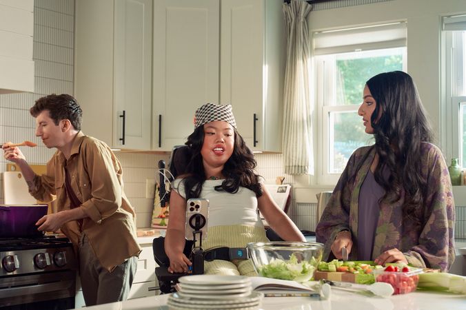 A SouthEast Asian woman with curled black hair seated in her power wheelchair gives her roommate instructions on how to prepare a salad in their bright kitchen. Her smart phone is mounted to her wheelchair. Behind them, a white male with Moebius Syndrome and a face that is paralyzed holds a wooden spoon up to his lips to taste a soup on the stove. The SouthEast Asian woman is wearing a white top with striped, yellow linen pants and a checkered headscarf. He has short, wavy, reddish brown hair and is wearing a tan button down shirt with the sleeves rolled up, showing arm tattoos. Their roommate has long dark hair and is wearing a purple floral dress.