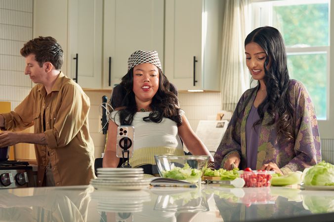 A SouthEast Asian woman with curled black hair seated in her power wheelchair watches her roommate prepare a salad in their bright kitchen. Her smart phone is mounted to her wheelchair. Behind them, a white male with Moebius Syndrome stirs a pot on the stove. The SouthEast Asian woman is wearing a white top with striped, yellow linen pants and a checkered headscarf. Her roommate has long dark hair and is wearing a purple floral dress.