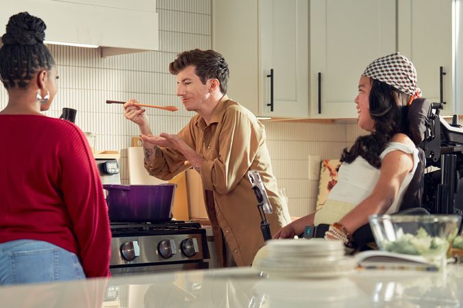 A white male with Moebius Syndrome and a face that is paralyzed cooks in a bright kitchen with his two roommates. He is standing over a stove with a purple pot, holding a spoon to his mouth, about to taste a sauce. Both roommates face him, watching the taste test. One roommate is a tan SouthEast Asian woman seated in her power wheelchair; the other is a black female. The male has short, wavy, reddish-brown hair and blue eyes. He is wearing a tan button-down shirt with the sleeves rolled up, showing arm tattoos. The SouthEast Asian woman has curled, black hair and is wearing a white top with striped, yellow linen pants, and a checkered head scarf. Her smart phone is mounted to her wheelchair.