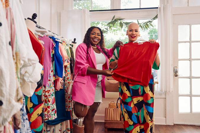 A bald African American person holds a black patterned skirt to her hips, deciding how it would look on her. Her friend, a Black woman with a prosthetic leg, holds up a red shirt to her friend's torso, seeing how it would match the patterned skirt. The two friends are in a boutique. The Black woman with a prosthetic leg has long, black curly hair and is wearing a vibrant pink blazer, and jean cut off shorts. The bald person is wearing a white and green striped crochet outfit and pointed white shoes. Clothing racks filled with high end garments are next to the women.