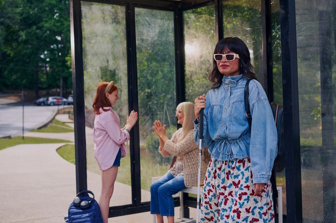 An Asian Filipino woman with Retinitis Pigmentosa and Usher Syndrome stands at a covered bus stop. She is wearing white sunglasses and is holding her white cane. She has on a jean jacket, a butterfly patterned skirt, and a black backpack. Behind her, a white European girl with red hair plays pattycake hand game with her grandmother, who is sitting on the bus stop bench. Behind the bus stop are several large trees and walking paths.