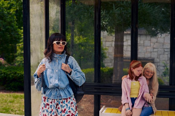 An Asian Filipino woman with Retinitis Pigmentosa and Usher Syndrome stands at a covered bus stop. She is wearing white sunglasses and is holding her white cane with her tattooed hands. She has on a jean jacket, a butterfly patterned skirt, and a black backpack. Behind her, a white European girl with red hair sits on her grandmother's lap on the bus stop bench and shows her a bracelet she is wearing. Behind the bus stop are several large trees, a landscaped garden, and a stone building.