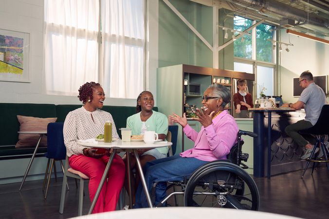 An African American mother, daughter, and auntie are smiling and chatting at a table in a modern cafe. The auntie is paraplegic and is seated in a manual wheelchair. She is smiling wide and raises her arms in excitement. She has short gray hair and is wearing a lavender silk blouse and dark blue jeans. The mother has short tightly curled red hair and is wearing a patterned blouse and red pants. The daughter is wearing a cochlear implant and has her braided hair pulled back into a pony tail. She is wearing a light green cardigan and jean shorts. Behind them, a barista prepares drinks and a man types on a laptop.