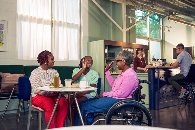 An African American daughter and auntie give each other a high five at a table in a modern cafe, while the girl's mother smiles and watches. The auntie is paraplegic and is seated in a manual wheelchair.  She has short gray hair and is wearing a lavender silk blouse and dark blue jeans. The mother has short tightly curled red hair and is wearing a patterned blouse and red pants. The daughter is wearing a cochlear implant and has her braided hair pulled back into a pony tail. She is wearing a light green cardigan and jean shorts. Behind them, a barista prepares drinks and a man types on a laptop.