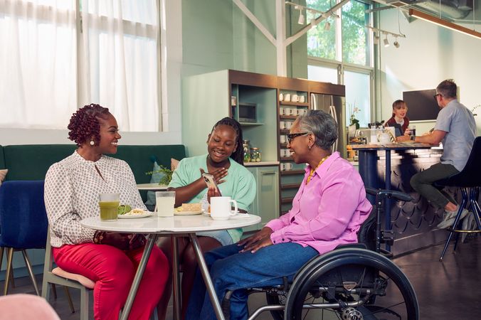 An African American girl shows her auntie something on her phone at a table in a modern cafe, while the girl's mother smiles broadly and watches. The auntie is paraplegic and is seated in a manual wheelchair. She has short gray hair is wearing a lavender silk blouse and dark blue jeans. The mother has short tightly curled red hair and is wearing a patterned blouse and red pants. The daughter is wearing a cochlear implant and has her braided hair pulled back into a pony tail. She is wearing a light green cardigan and jean shorts. Behind them, a barista hands a drink to a man seated the cafe bar.
