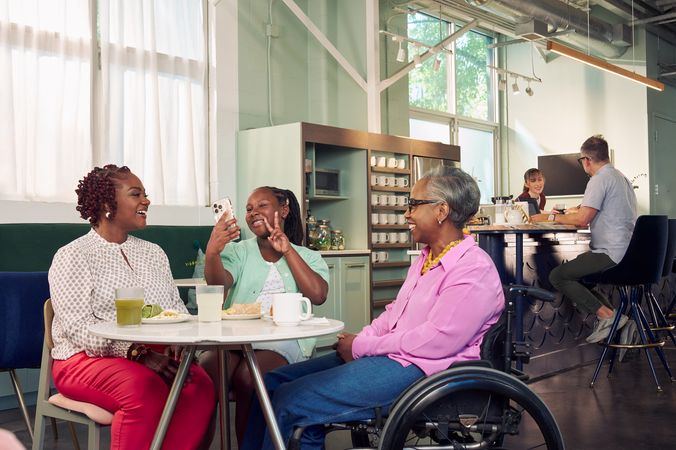 An African American girl takes a selfie and holds up two fingers in a peace sign in a modern cafe, while the girl's motherand auntie watch. The auntie is paraplegic and is seated in a manual wheelchair. She has short gray hair is wearing a lavender silk blouse and dark blue jeans. The mother has short tightly curled red hair and is wearing a patterned blouse and red pants. The daughter is wearing a cochlear implant and has her braided hair pulled back into a pony tail. She is wearing a light green cardigan and jean shorts. Behind them, a barista hands a drink to a man seated the cafe bar.