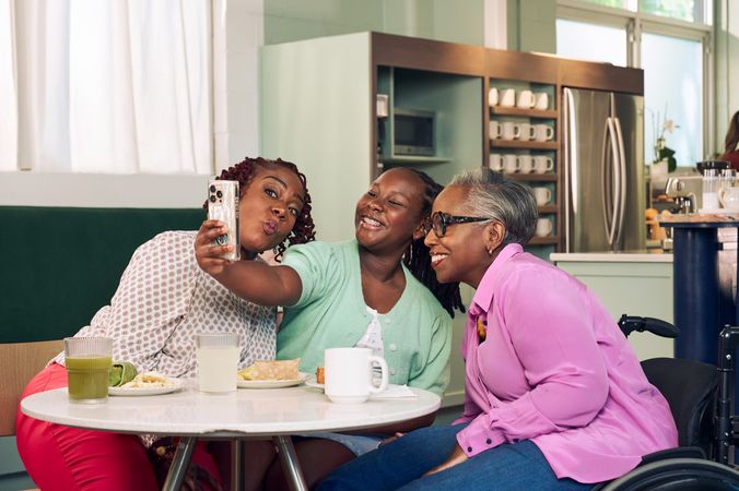 An African American girl takes a selfie with her mother and auntie in a modern cafe. The auntie is paraplegic and is seated in a manual wheelchair. She has short gray hair is wearing a lavender silk blouse and dark blue jeans. The mother has short tightly curled red hair and is puckering her lips in a silly pose. She is wearing a patterned blouse and red pants. The daughter is wearing a cochlear implant and has her braided hair pulled back into a pony tail. She is wearing a light green cardigan and jean shorts.