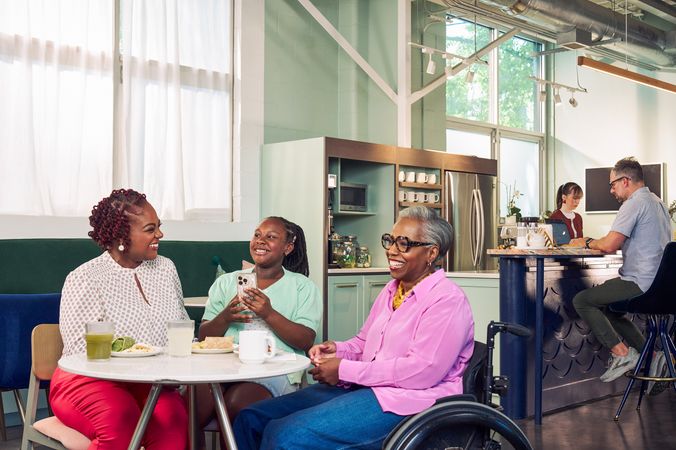 An African American girl shows smiles and holds her phone at a table in a modern cafe, while the girl's mother and auntie laugh together. The auntie is paraplegic and is seated in a manual wheelchair. She has short gray hair is wearing a lavender silk blouse and dark blue jeans. The mother has short tightly curled red hair and is wearing a patterned blouse and red pants. The daughter is wearing a cochlear implant and has her braided hair pulled back into a pony tail. She is wearing a light green cardigan and jean shorts. Behind them, a barista makes drinks and a man types on a laptop.