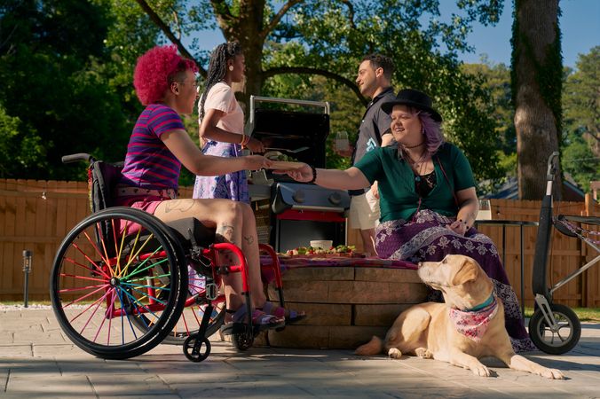 At a backyard barbeque, a white nonbinary person sits on a low stone table and passes a plate of food to a Black nonbinary person seated in a manual wheelchair with rainbow spokes. The white nonbinary person has a uniquely shapped dark gray rollator decorated with purple acrylic chains nearby. They have layered purple hair with two thin long braids on each side and are wearing a black hat, a green shirt, and a purple patterned skirt. The Black nonbinary person seated in their manual wheelchair has a curly pink fro with loc bangs and is wearing a purple and pink striped crop top, pink shorts, and purple sandals. They have several tattoos on both legs. A golden labrador retriever wearing a pink hankerchief looks up at the plate of food. Behind them, two friends chat at a grill. One friend is an African American woman and is holding a spatula. The other friend is a multi-ethnic man with curly black hair.