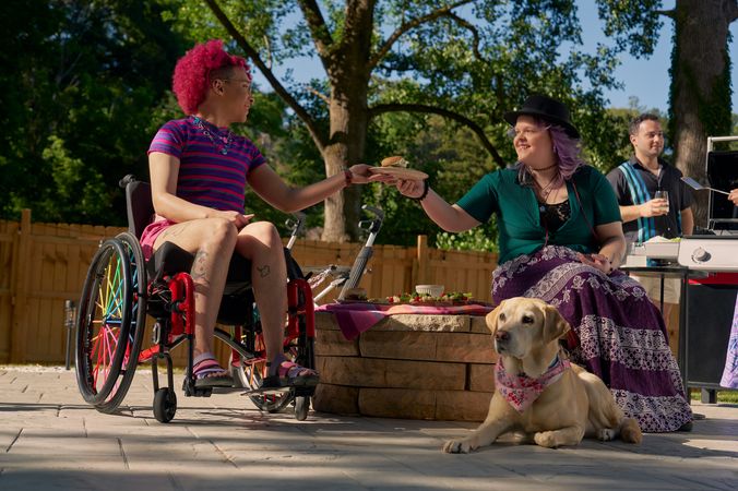 At a backyard barbeque, a white nonbinary person sits on a low stone table and passes a plate of food to a Black nonbinary person seated in a manual wheelchair with rainbow spokes. The white nonbinary person has a uniquely shapped dark gray rollator decorated with purple acrylic chains nearby. They have layered purple hair with two thin long braids on each side and are wearing a black hat, a green shirt, and a purple patterned skirt. The Black nonbinary person seated in their manual wheelchair has a curly pink fro with loc bangs and is wearing a purple and pink striped crop top, pink shorts, and purple sandals. They have several tattoos on both legs. A golden labrador retriever wearing a pink hankerchief sits at their feet. Behind them, another friend, a multi-ethnic man with curly black hair, holds a drink and chats near a grill.