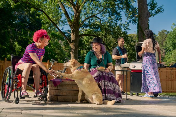 At a backyard barbeque, a golden labrador retriever places her paw in the palm of a Black nonbinary person's hand. The Black nonbinary person is seated in a manual wheelchair with rainbow spokes and has their mouth open in praise of the dog. Next to them, a white nonbinary person sits on a low stone table and holds a plate of food. They have layered purple hair with two thin long braids on each side and are wearing a black hat, a green shirt, and a purple patterned skirt. The Black nonbinary person seated in their manual wheelchair has a curly pink fro with loc bangs and is wearing a purple and pink striped crop top, pink shorts, and purple sandals. They have several tattoos on both legs. Behind them, two friends chat at a grill. One friend is an African American woman and is holding a spatula. The other friend is a multi-ethnic man with curly black hair.