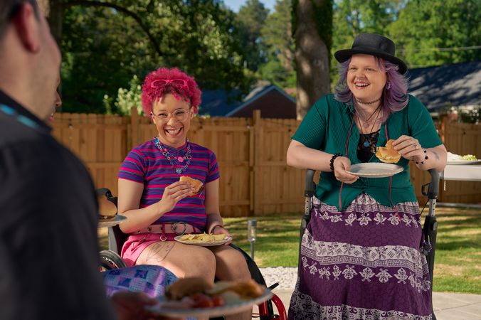 At a backyard barbeque, two nonbinary people sit in their mobility aids, laughing and holding plates of food. One nonbinary person, a Black person with a curly pink fro with loc bangs wearing glasses is seated in a manual wheelchair. They are wearing bright pink sunglassese on their head, a purple and pink striped crop top, pink shorts, and purple sandals. They have several tattoos on both legs. The second nonbinary person, a white person with layered purple hair with two long thin red braids on each side, is seated on a dark gray rollator. They are wearing a black hat, a green shirt, and a purple patterned skirt.