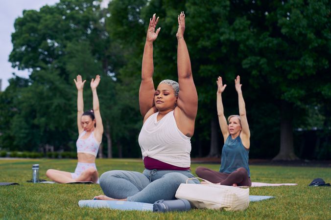 At an outdoor yoga class, a Black woman sits cross-legged with her eyes closed and hands extended straight overhead with palms facing inward toward each other. Her bag and water bottle are next to her. She has icy blonde shortly cut hair and is wearing a white and red layered tank and gray drawstring pants. Behind her, two tall women sit on their mats in the same pose. The class is in a grassy park with many large trees.