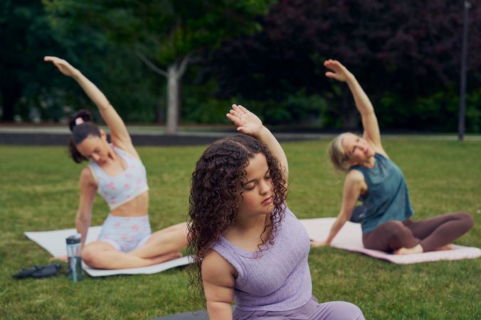 At an outdoor yoga class, a young white woman with dwarfism sits in side bend stretch, cross-legged with her eyes closed, one hand resting on the ground while the opposite arm reaches up and arches over to the side. She has long, curly brown hair and is wearing a purple ribbed tank with comfortable, loose purple pants. Behind her, two tall women sit on their mats in the same pose. The class is in a grassy park with many large trees.