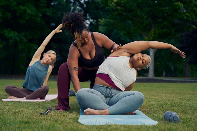 A Black woman yoga instructor supports a Black woman student move into a modified side bend stretch. The student sits in a cross-legged position, leaning to her right side with her left arm reaching over to stretch her left side. The instructor stands behind her, gently placing one hand on her outstretched arm, and the other on her upper thigh. The student has a walking cane placed on the grass next to her yoga mat. She has icy blonde shortly cut hair and is wearing a white and red layered tank and gray drawstring pants. The instructor has her black hair pulled back into a natural ponytail with dyed bangs and is wearing a black tank and maroon yoga pants. Behind her, another woman sits in the same pose, unsupported.