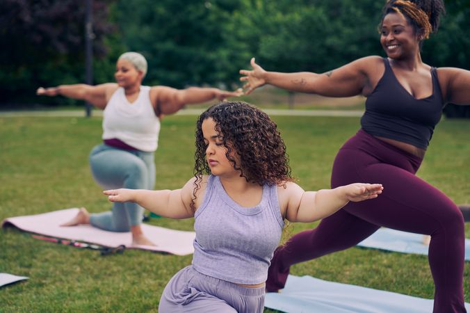 At an outdoor yoga class, a young white woman with dwarfism stands in Warrior pose, with her eyes closed, both arms extended out parallel to the ground, one facing forward and one reaching back. She has long, curly brown hair and is wearing a purple ribbed tank with comfortable, loose purple pants. Behind her, a tall Black woman instructor smiles while also in the same pose. Another Black woman is in a modified form of the pose, kneeling. Her cane is next to her mat. The class is in a grassy park with many large trees.