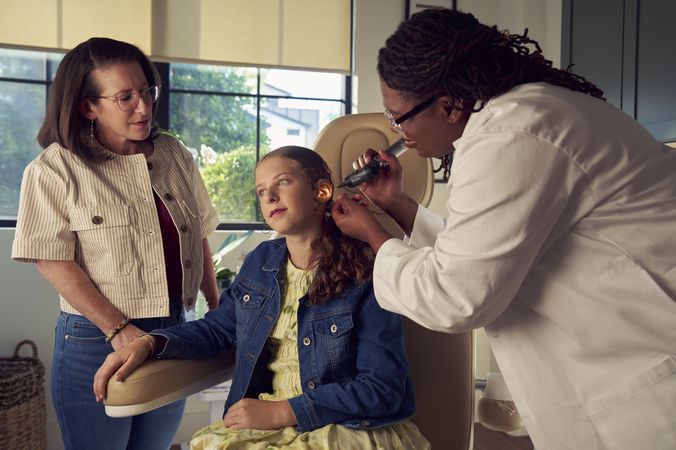A Black deafBlind audiologist with a cochlear implant and glasses smiles and uses an otoscope to look into the ears of a young white deaf teen with braces seated in a medical treatment chair. The teen is softly smiling and has long, wavy sandy blonde hair and is wearing a jean jacket, and a yellow blouse. The teen's mother is standing next to her, observing the audiologist use the tool.