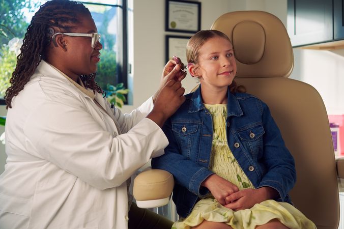 A Black deafBlind audiologist with a cochlear implant and glasses places a cochlear implant on a young white deaf teen with braces seated in a medical treatment chair. The teen is smiling and has long, wavy sandy blonde hair and is wearing a jean jacket, and a yellow blouse and skirt. Framed professional degrees are hanging on the wall behind them.