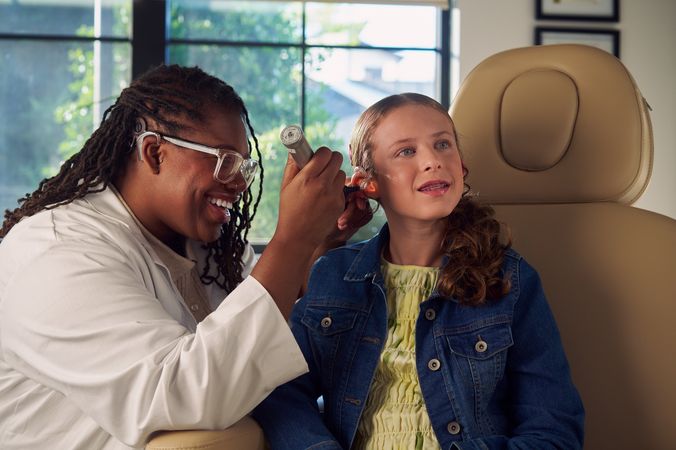 A Black deafBlind audiologist with a cochlear implant and glasses smiles and uses an otoscope to look into the ears of a young white deaf teen with braces seated in a medical treatment chair. The teen is smiling and has long, wavy sandy blonde hair and is wearing a jean jacket, and a yellow blouse and skirt. Framed professional degrees are hanging on the wall behind them.
