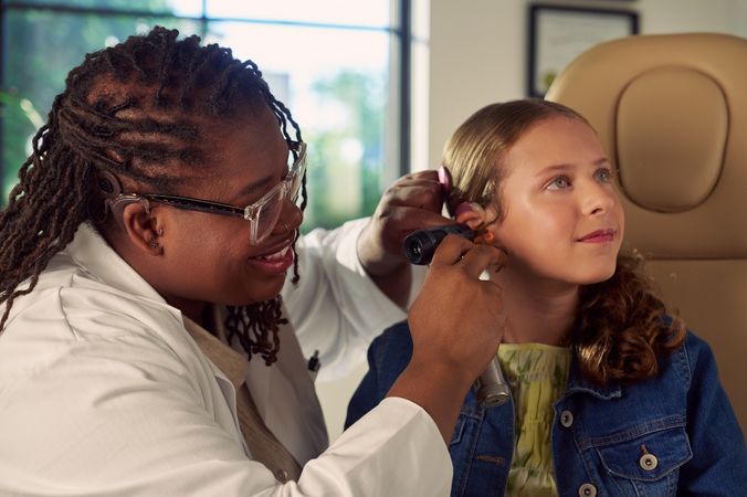 A Black deafBlind audiologist with a cochlear implant and glasses smiles and uses an otoscope to look into the ears of a young white deaf teen with braces seated in a medical treatment chair. The teen is softly smiling and has long, wavy sandy blonde hair and is wearing a jean jacket, and a yellow blouse.