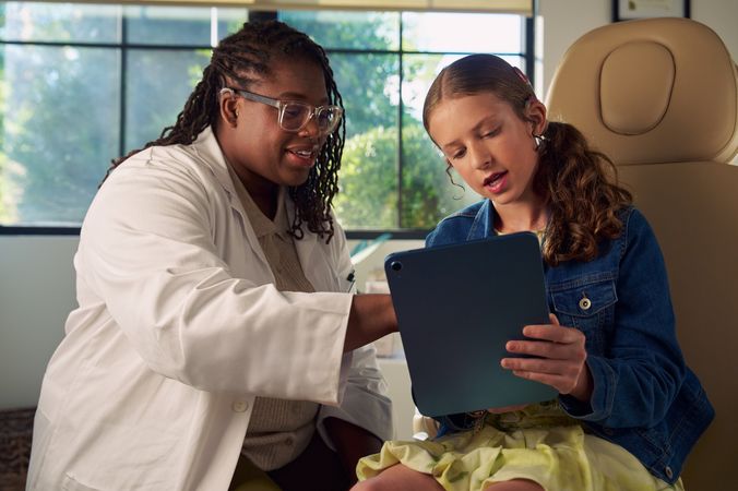 A Black deafBlind audiologist with a cochlear implant and glasses points to something visible on a tablet that a young white deaf teen with braces is holding. The teen is seated in a medical treatment chair and looks at the tablet with curiosity. She and has long, wavy sandy blonde hair and is wearing a jean jacket, and a yellow blouse and skirt.