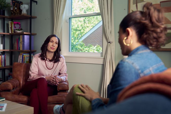 An Eastern European white woman therapist with a facial scar sits in a large chair and looks at a client seated on a couch in front of her. The client is a Latina woman with dark brown wavy hair pulled into a ponytail. The therapist's office has several books on display, an abstract art piece, and a window letting in natural light. The therapist is wearing a silk pink top, maroon dress pants, and a minimalist necklace. The client is wearing gold hoop earrings, a jean jacket, green skirt, and sneakers.