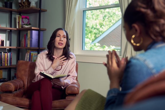 An Eastern European white woman therapist with a facial scar  sits in a large chair and looks at a client seated on a couch in front of her. She is holding a pen and open notebook. The client is a Latina woman with dark brown wavy hair pulled into a ponytail. The therapist's office has several books on display and a window letting in natural light. The therapist is wearing a silk pink top, maroon dress pants, and a minimalist necklace. The client is wearing gold hoop earrings, a jean jacket, and green skirt.