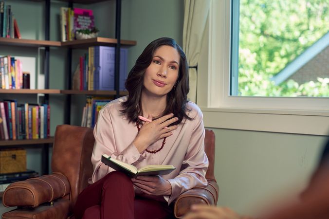 An Eastern European white woman therapist with a facial scar sits in a large chair and holds her hand up to her chest with an understanding look on her face. She is holding a pen and open notebook, and is looking at a client off camera. The therapist's office has several books on display and a window letting in natural light. The therapist is wearing a silk pink top, maroon dress pants, and a minimalist necklace.
