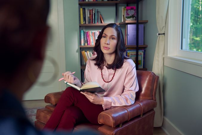An Eastern European white woman therapist with a facial scar  sits in a large chair with an understanding look on her face. She is holding a pen and open notebook, and is looking at a client off camera. The therapist's office has several books on display and a window letting in natural light. The therapist is wearing a silk pink top, maroon dress pants, and a minimalist necklace.