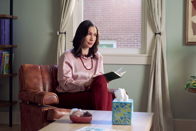 An Eastern European white woman therapist with a facial scar sits in a large chair looks at a client off camera. She is holding an open notebook. A tissue box and a red marble heasrt-shaped bowl of crystals are on a low coffee table in front of her. The therapist's office has several books on display and a window letting in natural light. The therapist is wearing a silk pink top, maroon dress pants, and a minimalist necklace.