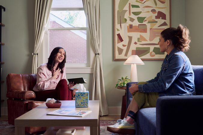 An Eastern European white woman therapist with a facial scar  sits in a large chair and looks at a client seated on a couch in front of her. She is holding her hand to her chest and smiles at her client knowingly. The client is a Latina woman with dark brown wavy hair pulled into a ponytail. A tissue box, a red marble heasrt-shaped bowl of crystals, and a magazine are on a low coffee table in the center of the office. An abstract art piece, a lamp, and a small house plant are next and a window letting in natural light. The therapist is wearing a silk pink top, maroon dress pants, and a minimalist necklace. The client is wearing gold hoop earrings, a jean jacket, green skirt, and sneakers.