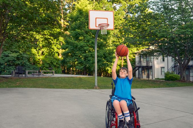 A white teen boy seated in a manual wheelchair holds a basketball over his head, with a look of concentration on his face. He is playing at a basketball court at a neighborhood park. He has short brown hair and is wearing blue athletic wear.